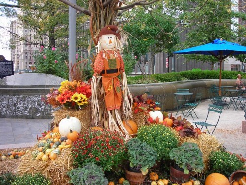 A festive scarecrow surrounded by colorful flowers, pumpkins, and hay in a park setting.