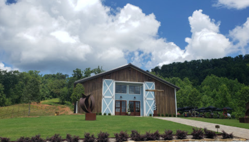 A rustic barn with white doors, surrounded by green trees and a blue sky with fluffy clouds.