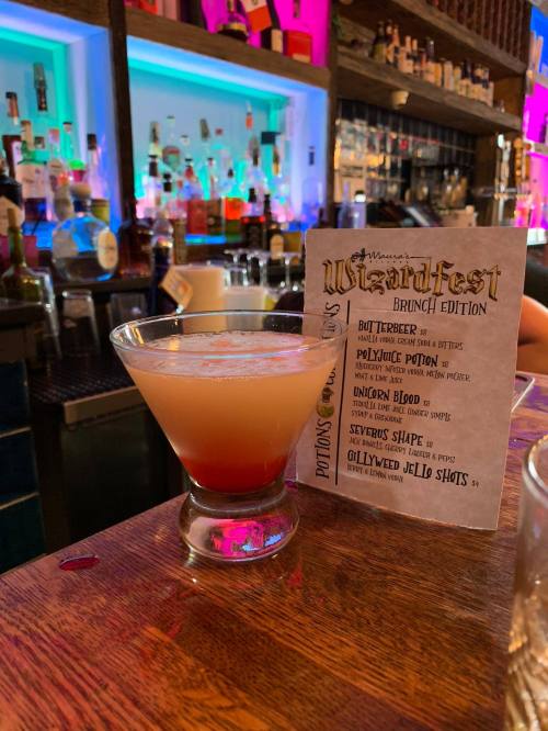 A colorful cocktail in a martini glass sits on a bar, next to a menu for a themed brunch event.
