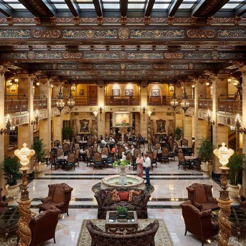 Luxurious hotel lobby with ornate decor, elegant seating, and guests mingling under a glass ceiling.