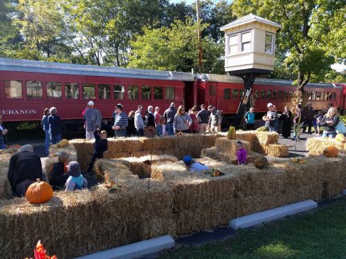 A crowd gathers around a hay maze with pumpkins, near a vintage red train and a lookout tower in a park setting.