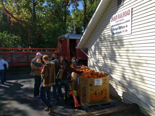 A group of people gathers near a ticket office, with a box of pumpkins outside and a train car in the background.