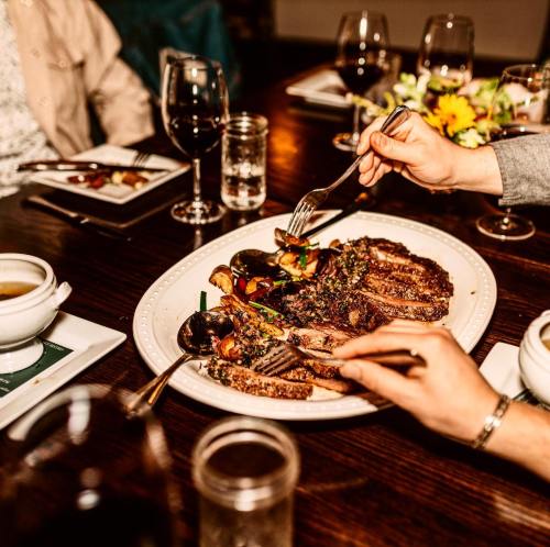 A hand reaches for a platter of sliced meat with colorful vegetables, surrounded by wine glasses and plates on a dining table.