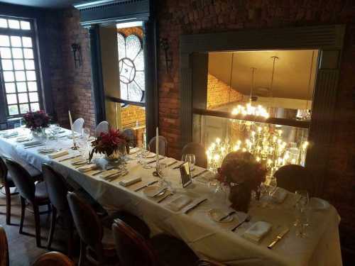 Elegant dining setup with a long table, floral centerpieces, and soft lighting in a cozy, brick-walled room.