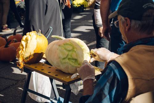 A person carves a large pumpkin on a table at a market, surrounded by other pumpkins and people.