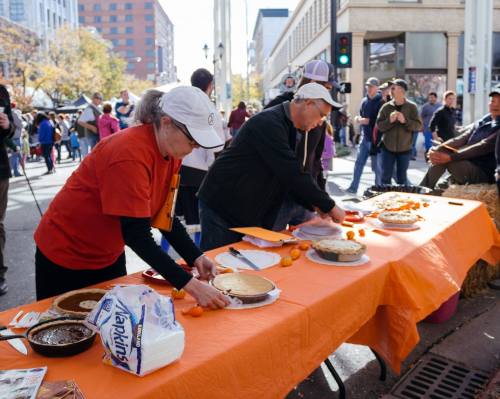 Two people prepare pies on an orange table at a busy outdoor event, with a crowd and buildings in the background.