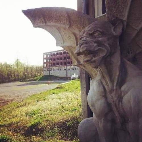 A stone gargoyle with bat-like wings stands near an abandoned building, surrounded by greenery and a clear sky.