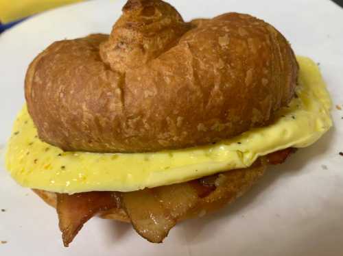 A croissant sandwich with a fluffy egg patty and crispy bacon, served on a white plate.