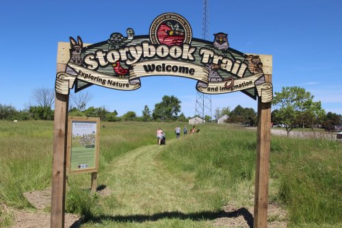 A wooden sign welcomes visitors to the Storybook Trail, surrounded by green grass and trees under a clear blue sky.