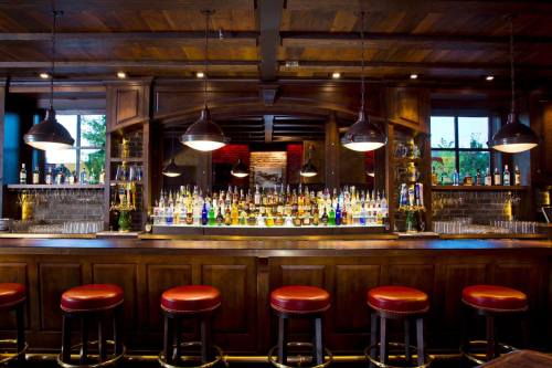 A wooden bar with colorful bottles, illuminated by pendant lights, and red leather stools in a cozy setting.