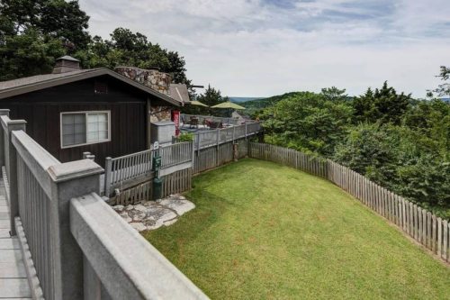 A scenic view of a house with a fenced yard, surrounded by trees and hills under a cloudy sky.