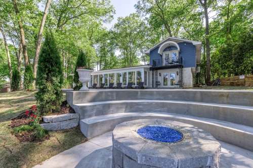 A modern home surrounded by trees, featuring a stone patio and a fire pit with blue glass stones.