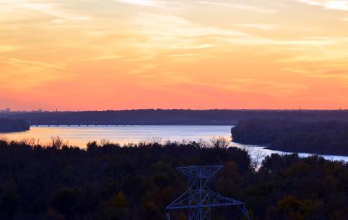 A serene sunset over a river, with silhouettes of trees and a power line in the foreground.