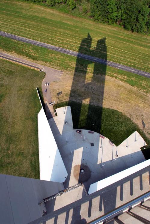 Aerial view of a building casting a long shadow on the ground, with green fields and a pathway in the background.