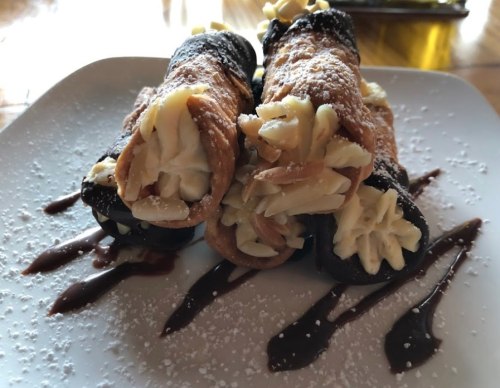Three cannoli filled with cream, dusted with powdered sugar, and drizzled with chocolate sauce on a white plate.