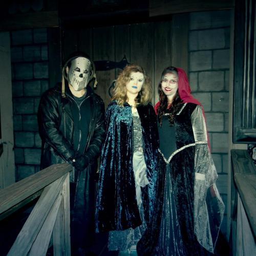 Three people in costumes stand in a dimly lit setting, with one wearing a mask and the others in cloaks and makeup.
