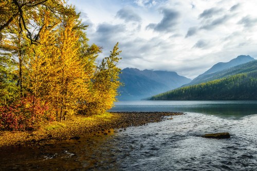 A serene lake scene with autumn trees, mountains in the background, and a cloudy sky reflecting on the water.