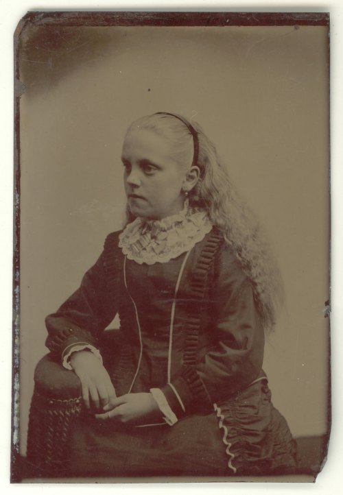 A young girl with long, wavy hair sits in profile, wearing a vintage dress with a ruffled collar and a serious expression.