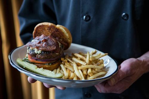 A chef holds a plate with a burger topped with bacon, onions, and pickles, alongside a serving of French fries.