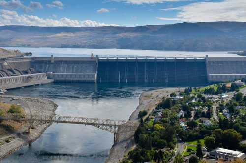 A large dam spans a river, with mountains in the background and a small town visible nearby. Clear blue sky above.