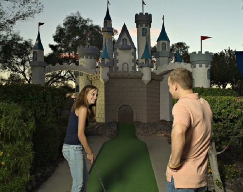 A young couple smiles at each other in front of a whimsical castle-themed mini-golf course at sunset.