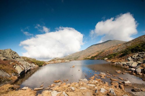 A serene landscape featuring a calm lake surrounded by rocky terrain and mountains under a blue sky with fluffy clouds.