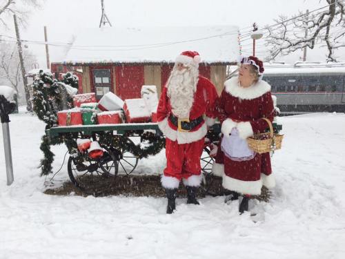 Santa and Mrs. Claus stand beside a festive wagon filled with gifts, surrounded by falling snow.