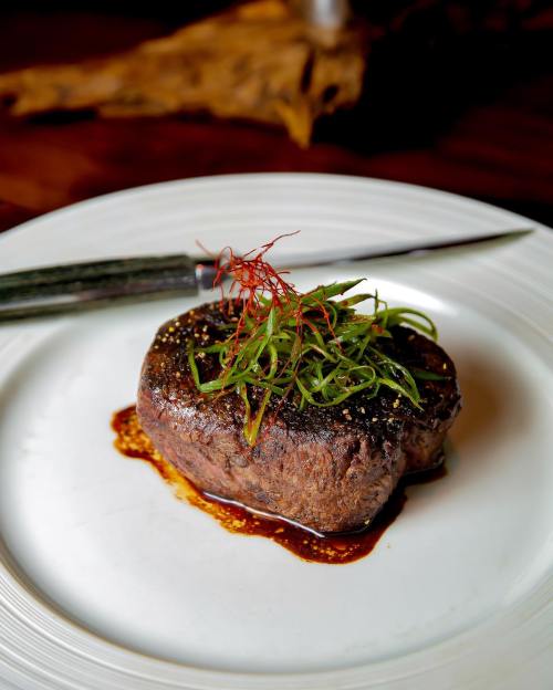A perfectly cooked steak garnished with green onions and red threads, served on a white plate with a knife beside it.