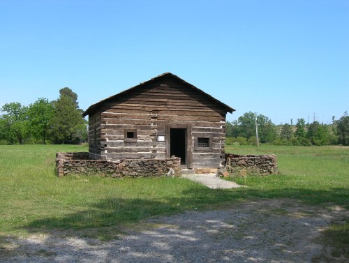 A rustic log cabin with a stone foundation, surrounded by green grass and trees under a clear blue sky.