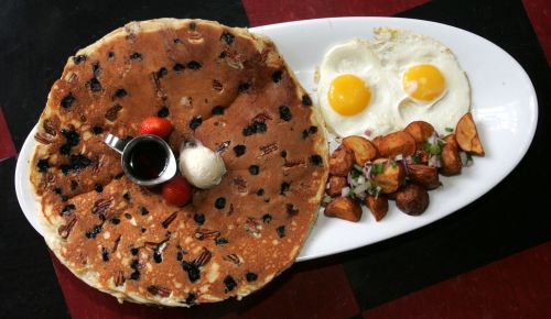 A large pancake topped with nuts and berries, served with two sunny-side-up eggs and seasoned potato wedges.