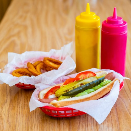 A hot dog with toppings and a side of curly fries, accompanied by yellow and pink condiment bottles on a wooden table.