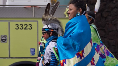 A woman in a blue shawl and a child in traditional attire smile during a cultural event, with a green vehicle in the background.