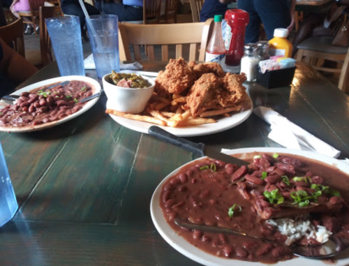 A table with two plates of red beans and rice, fried chicken, fries, and a side of greens, with drinks in the background.