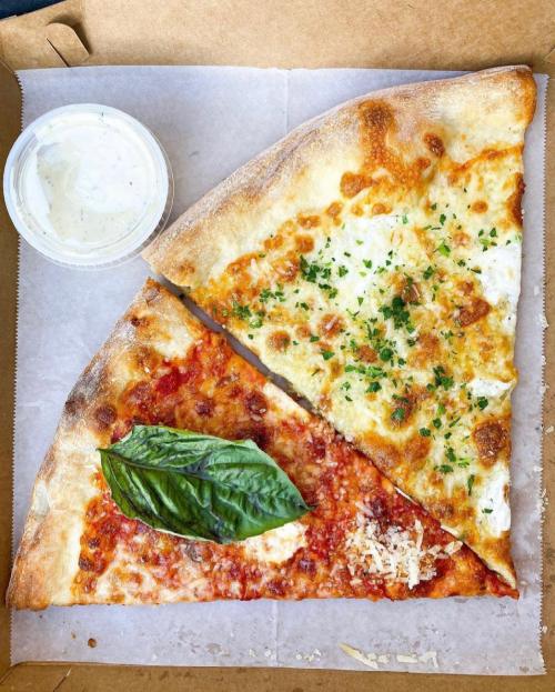 A pizza box containing two slices: one with tomato sauce and basil, the other with cheese and herbs, alongside a dipping sauce.