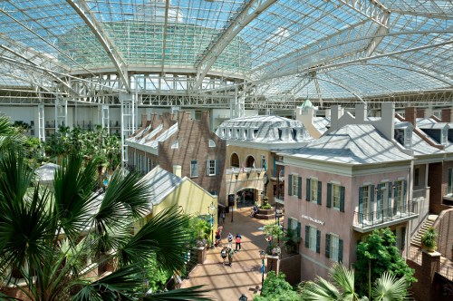 A spacious indoor atrium with a glass roof, featuring shops, greenery, and visitors walking along pathways.