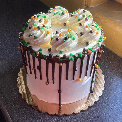 A round cake with white frosting, chocolate drizzle, and colorful decorations on top, placed on a gold cake board.