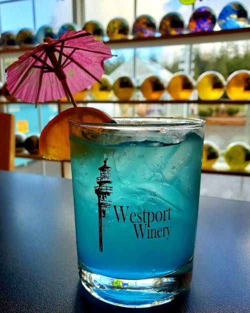 A blue cocktail with a pink umbrella and lemon slice, featuring the Westport Winery logo, set against colorful glass decor.