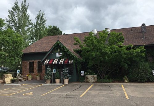 A brick building with a sloped roof, surrounded by greenery and planters, featuring a striped awning at the entrance.