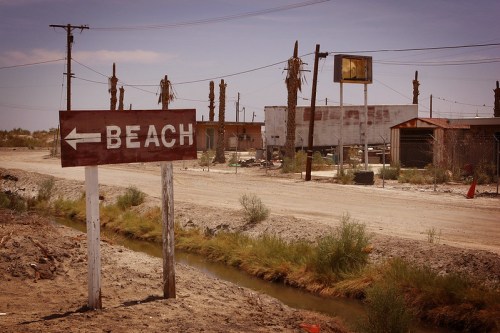 A weathered sign pointing left to a beach, with a desolate road and abandoned buildings in the background.