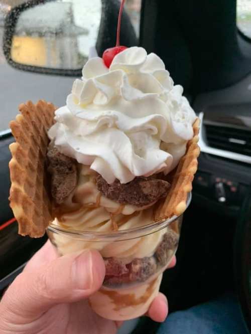 A hand holding a dessert cup filled with ice cream, whipped cream, caramel, cookie pieces, and topped with a cherry.