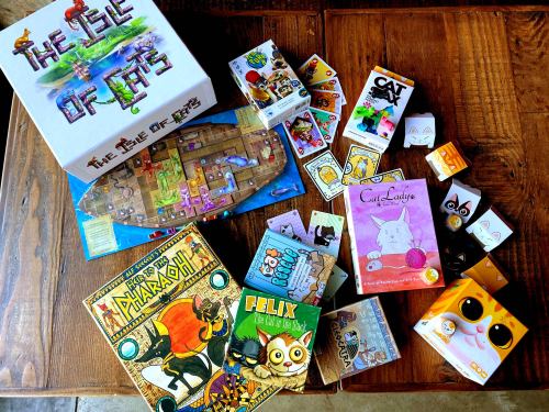 A collection of board games and card games featuring cats, spread out on a wooden table.