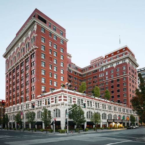 Historic red brick hotel with a modern addition, featuring large windows and trees lining the street corner.