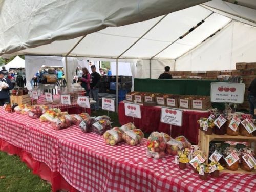 A market stall under a tent displays various fruits and apple butter, with prices labeled on signs.