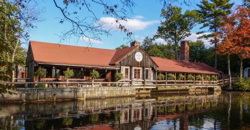 A rustic wooden building by a calm lake, surrounded by trees with autumn foliage and a clear blue sky.