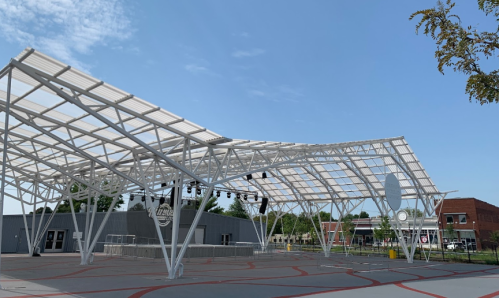 A modern outdoor stage with a large canopy, surrounded by a paved area and buildings in the background under a clear blue sky.