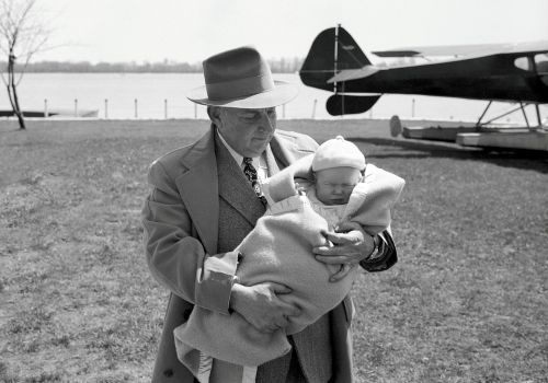 A man in a hat holds a wrapped baby outdoors near a plane, with a river in the background.