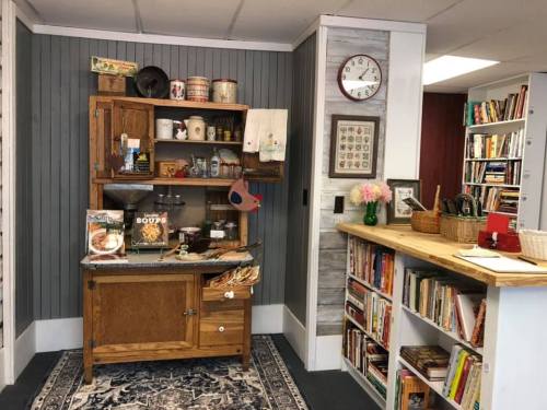 A cozy kitchen corner featuring a wooden shelf with decor, cookbooks, and a small table with flowers and a clock.