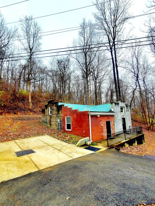 A small, brick building with a green roof, surrounded by bare trees and fallen leaves on a cloudy day.