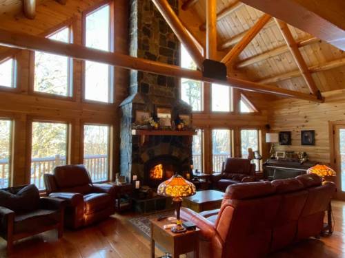 Cozy cabin interior with a stone fireplace, wooden beams, and large windows, featuring comfortable leather seating.