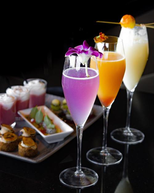 Three colorful cocktails in tall glasses, garnished with flowers and fruit, alongside a platter of assorted appetizers.